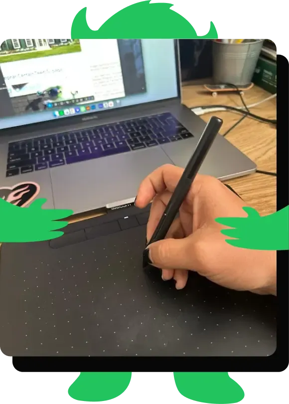 A hand drawing on a graphics tablet beside a laptop, framed inside a green Beautiful Beast silhouette